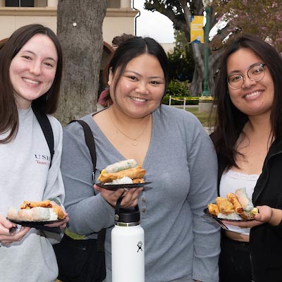 Three Fullerton College students smile at the camera with snacks in their hands.