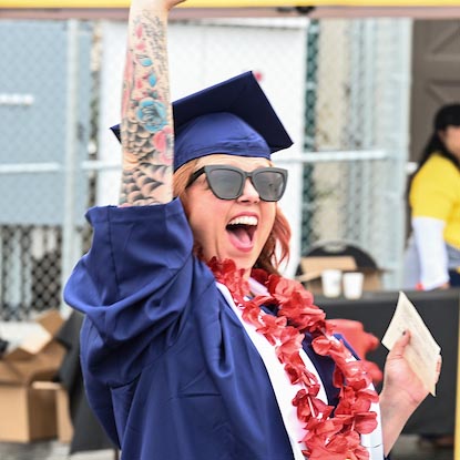 A woman in regalia smiles with her arm in the air during a Fullerton College graduation ceremony.